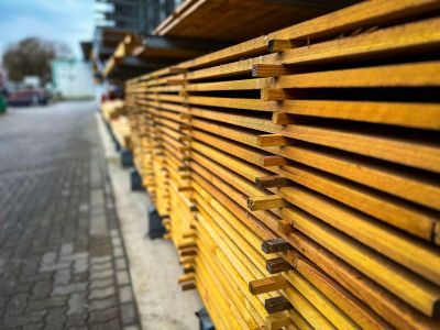 Gestapelte Holzbretter lagern drau&szlig;en entlang gepflastertem Weg, zeigen geordnetes Baumaterial.